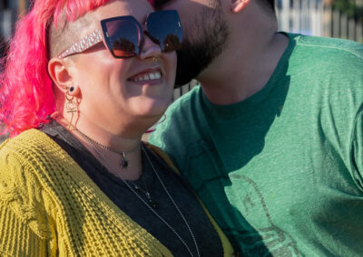 A slightly out of focus building in the background. Man in the background has short dark hair and a dark beard and is kissing his partner on the temple. In the foreground is his partner, with shoulder length bright pink colored hair wearing sunglasses with a smile.