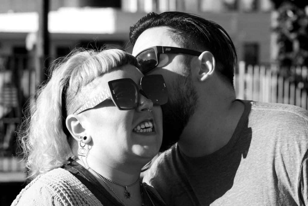 Black and white image. With a slightly out of focus building in the background. Man in the background has short dark hair and a dark beard and is kissing his partner on the temple. In the foreground is his partner, with shoulder length light colored hair wearing sunglasses with a playful grimace.