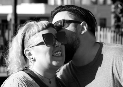 Black and white image. With a slightly out of focus building in the background. Man in the background has short dark hair and a dark beard and is kissing his partner on the temple. In the foreground is his partner, with shoulder length light colored hair wearing sunglasses with a playful grimace.