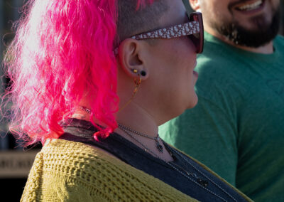In front is someone with shoulder length bright pink hair and sunglasses, looking back at their partner. In the back their partner has short dark hair and a short beard and is looking up at their partner with a smile.