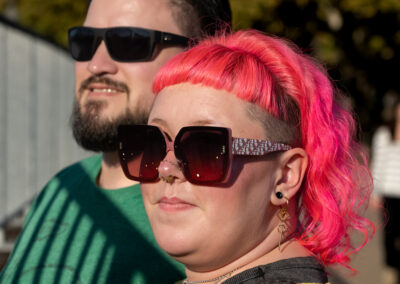 Two people standing together next to a barred railing fence. In the back is someone with short dark hair and a short beard wearing sunglasses and looking off to the left. In front of them is someone with shoulder-length bright pink hair, also wearing sunglasses and looking off to the left.