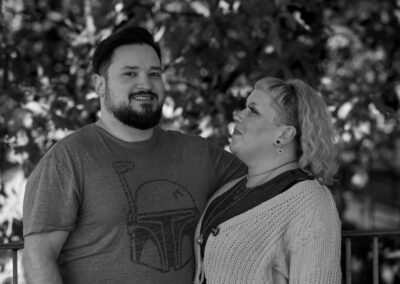 Black and white image of a couple against the backdrop of slightly out of focus foliage from a tree. The partner on the left has short, dark hair and a beard and is looking towards the camera. The partner on the right is looking up at him and has shoulder-length, light colored hair.
