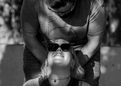 A dark-haired individual bending over the face of their partner with light hair. Their partner has sunglasses on, and is looking up at their partner smiling.