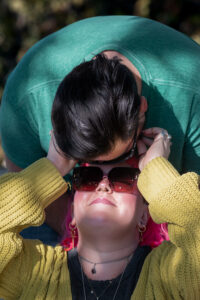 A dark-haired individual bending over the face of their partner with pink hair. Their partner has their hands up as they're looking up at them caressing their face with their fingers.