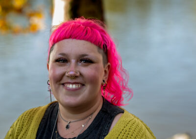 Background is an out of focus river with foliage from a tree. Pictured is a person with shoulder-length bright pink hair with a big smile.