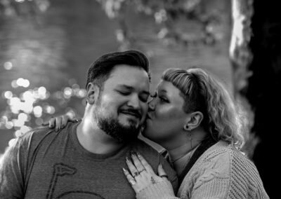 Black and white image. Background is a river with slightly out of focus foliage from a tree. Pictured is a couple, one with shoulder-length, light colored hair with one arm around the shoulder and the other hand on the other shoulder of their partner and kissing them on the cheek. Their partner is leaning in with short dark hair and a dark beard.
