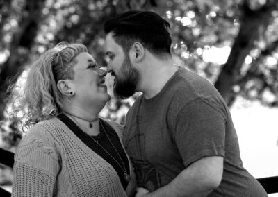 Black and white image. Background is slightly out of focus trees overhanging a river. There is a person with short dark hair and a dark beard leaning in toward their partner touching their nose with theirs. Their partner has shoulder-length light colored hair and is smiling as they touch noses with their partner.