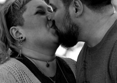 Black and white image. Background is out of focus trees. Pictured is a couple kissing, one with short dark hair and a beard, the other with shoulder-length, light colored hair.