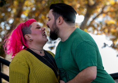 Background is slightly out of focus trees overhanging a river. There is a person with short dark hair and a dark beard leaning in toward their partner with their wide open. Their partner has shoulder-length bright pink colored hair and is looking up at their partner with a playful grimace. Slightly different from the prior images.