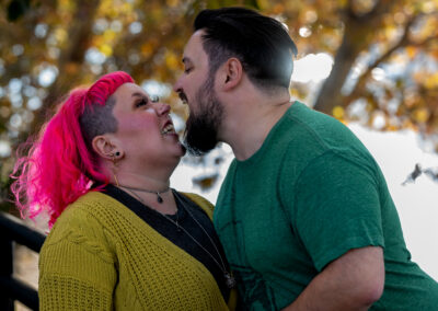 Background is slightly out of focus trees overhanging a river. There is a person with short dark hair and a dark beard leaning in toward their partner with their wide open. Their partner has shoulder-length bright pink colored hair and is looking up at their partner with a playful grimace.