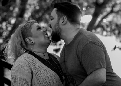 Black and white image. Background is slightly out of focus trees overhanging a river. There is a person with short dark hair and a dark beard leaning in toward their partner with their wide open. Their partner has shoulder-length light colored hair and is looking up at their partner with a playful grimace.