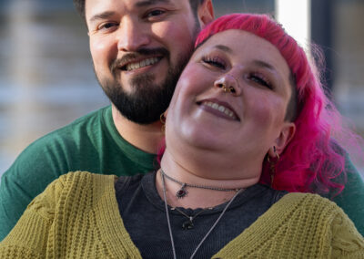 Background is a river. There is a person with short dark hair and a dark beard looking at the camera and smiling. The person in front of them has shoulder length bright pink hair and leaning back against them.