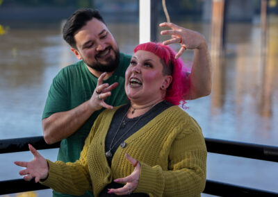 Background is a river. There is a person with short dark hair and a dark beard with his hands up looking like claws framing the face of the person in front of them. The person in front of them has shoulder length bright pink hair and is making a face.