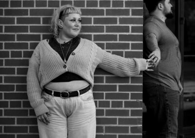 A black and white picture. The backdrop is the outside corner of a brick wall. A person with light colored hair, and a light colored shrug over a dark colored shirt and light colored jeans is leaning against the wall, looking to the right, with their right arm reaching out to grip the fingers of the person around the corner who we see in profile, with dark hair and a plain colored t shirt.