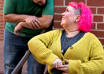 Background is a brick wall, with a short stair railing. There is a person with shoulder-length bright pink hair and a yellow-green shrug over a dark grey shirt and light washed jeans, leaning to the left against the railing. The person next to them has dark, short hair and a dark-colored beard, wearing a cool green t-shirt, and is leaning down onto the railing close to the other person. Both are looking at each other.
