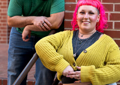 Background is a brick wall, with a short stair railing. There is a person with shoulder-length bright pink hair and a yellow-green shrug over a dark grey shirt and light washed jeans, leaning to the left against the railing. The person next to them has dark, short hair and a dark-colored beard, wearing a cool green t-shirt, and is leaning down onto the railing close to the other person. Both are looking straight at the camera.