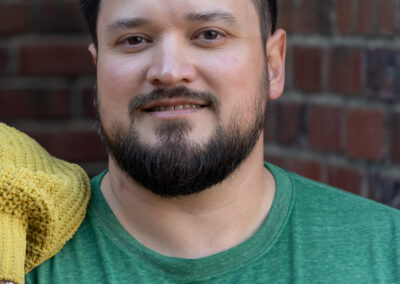 A close up of a man with dark hair and a dark beard, wearing a cool green shirt, against the backdrop of the inside corner of two brick walls.