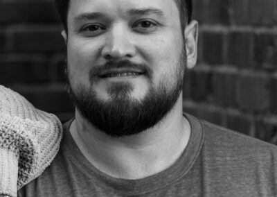 A black and white close up of a man with dark hair and a dark beard against the backdrop of the inside corner of two brick walls.