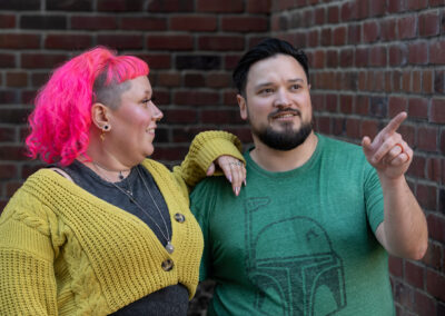 Background is the interior corner of two brick walls. There is a person with shoulder-length bright pink hair, and a yellow-green shrug over a dark grey shirt, with their arm up on the shoulder of a person next to them with dark, short hair and a dark-colored beard, wearing a cool green t-shirt. The person with pink hair is looking over at the person with dark hair, and the person with dark hair is looking slightly to the right and pointing.