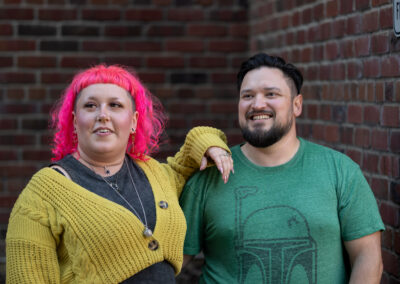 Background is the interior corner of two brick walls. There is a person with shoulder-length bright pink hair, and a yellow-green shrug over a dark grey shirt, with their arm up on the shoulder of a person next to them with dark, short hair and a dark-colored beard, wearing a cool green t-shirt. Both are looking off to the left and up slightly.