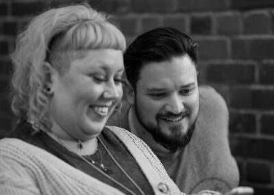 A black and white image. The background is brick, with a man slightly out of focus in the background with dark hair and a beard, leaning over to look at something. In the foreground is a woman with light colored hair, smiling, looking down at something in her hands.