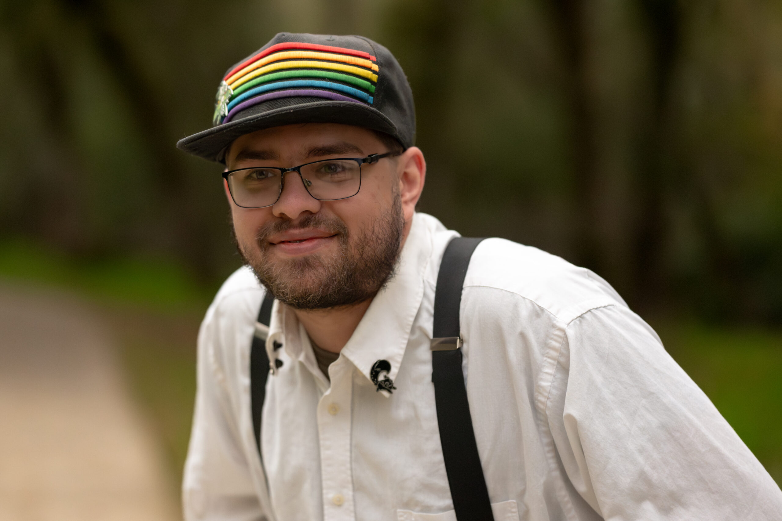 man wearing a baseball cap with rainbow stripes across the front, with glasses and a beard. He's wearing a white collared, button-down shirt with suspenders, and is set against an out of focus background of trees and foliage with a pat leading off to the left of the image.