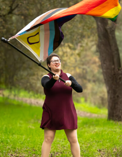 A person wearing a short maroon dress with black sleeves and black boots, with short dark hair and glasses, waving a progress pride flag overhead, looking up at it.