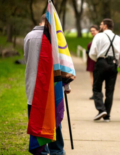 A group walking away from the camera, with the focus being on a young person carrying a progress pride flag over his shoulder.