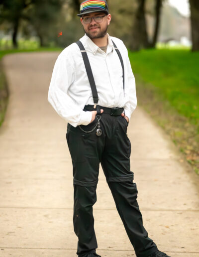 A young man wearing a dark colored hat with rainbow stripes across the front of it, wearing a white collared button down shirt with dark colored suspender straps and dark pants, with his hands in his pockets, looking at the camera.