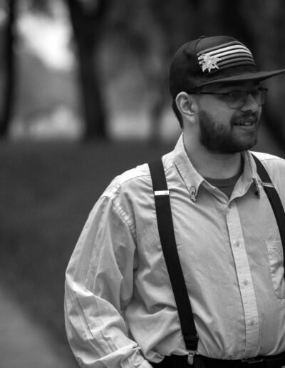 A black and white picture. A young man wearing a dark colored hat with stripes across the front of it, wearing a white collared button down shirt with dark colored straps with his hands in his pockets, looking off to the right.