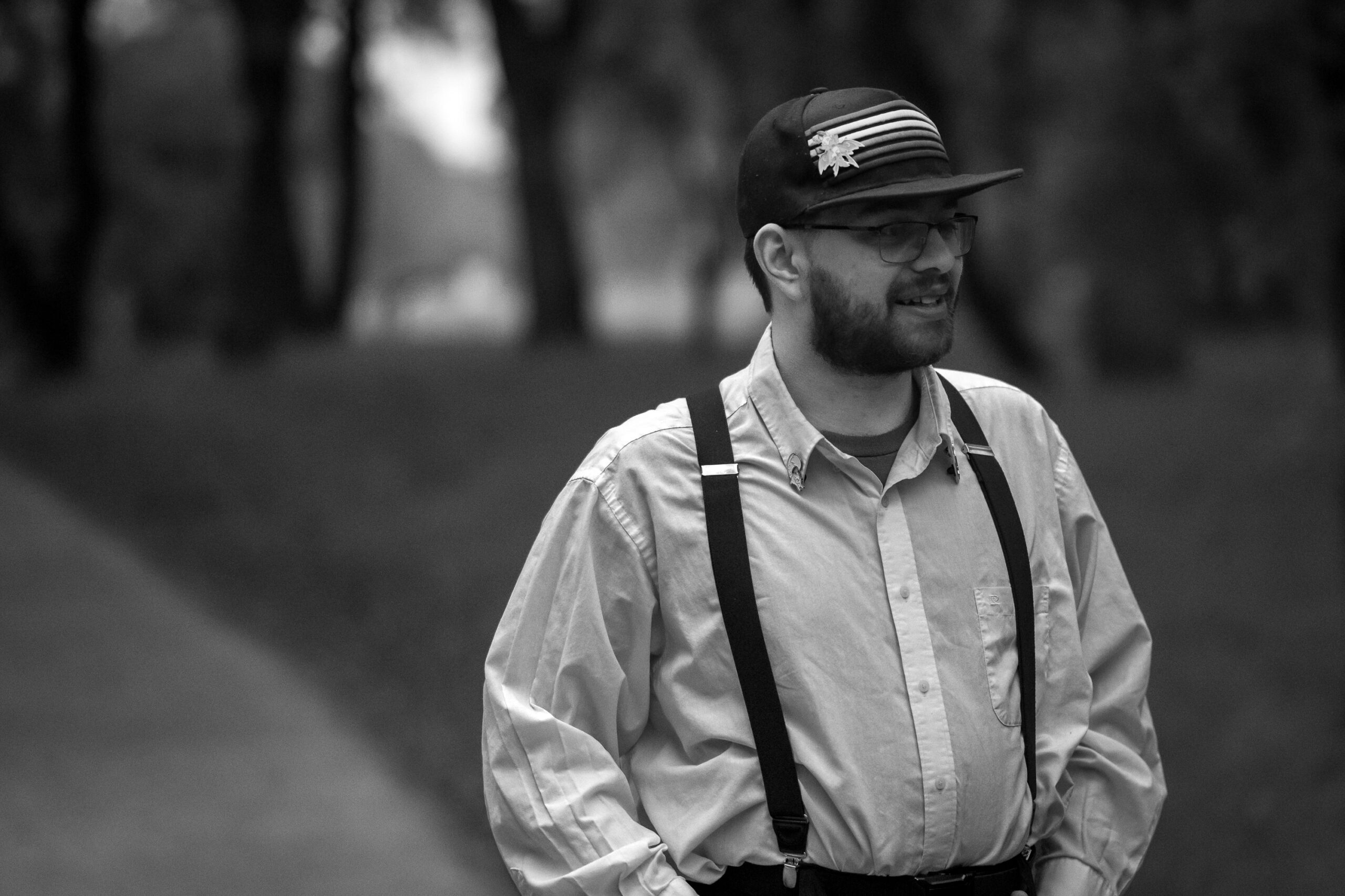 A black and white picture. A young man wearing a dark colored hat with stripes across the front of it, wearing a white collared button down shirt with dark colored straps with his hands in his pockets, looking off to the right.