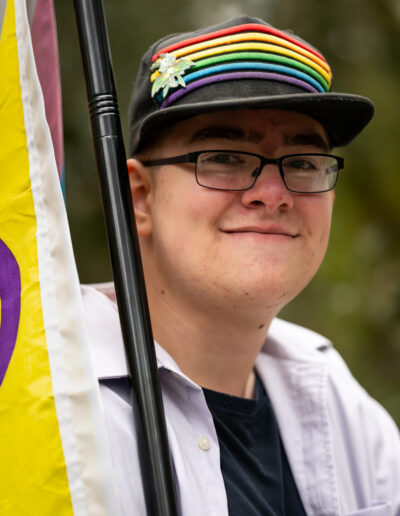 A young man wearing glasses with a dark colored baseball cap with rainbow stripes across the front. He has a flag tilting over one shoulder.