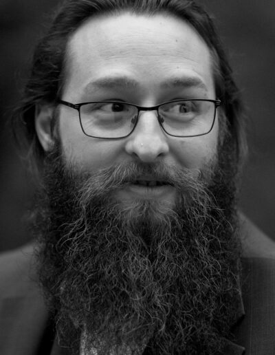 A black and white photo of a young man with dark hair and a longer, dark beard and glasses.