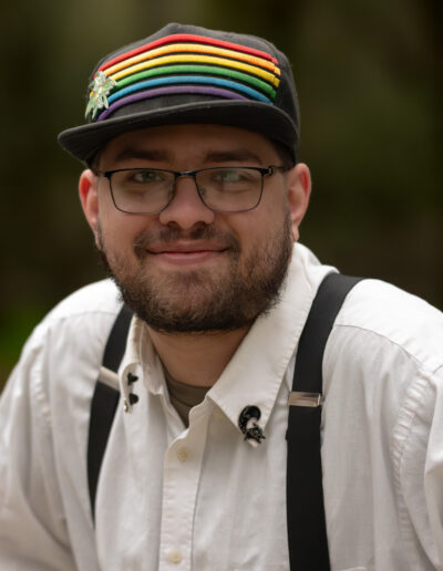 A young man wearing a dark colored baseball cap with rainbow stripes on it, wearing a white collared button down shirt with suspenders. He wears glasses and has a dark, short beard.
