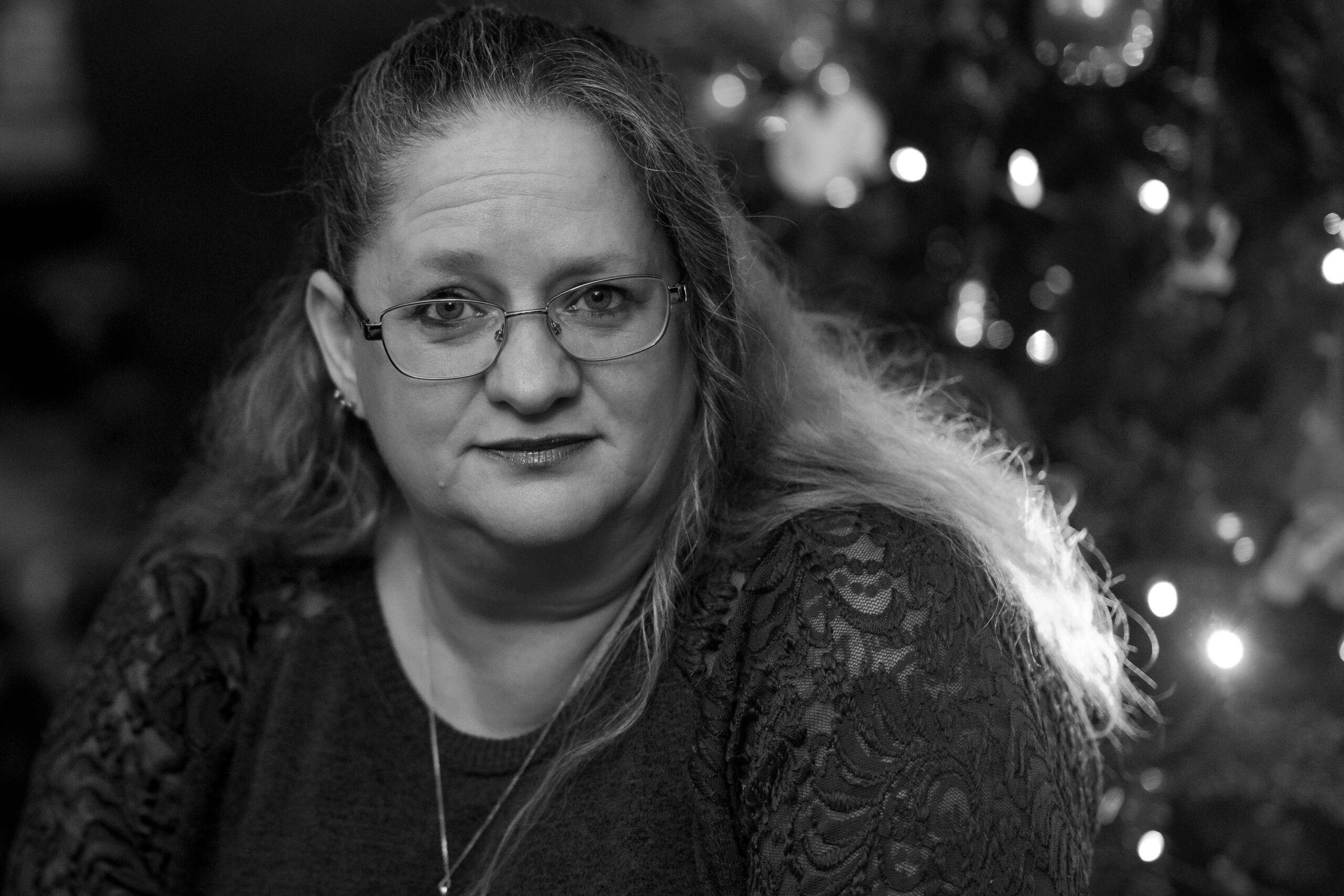 A black and white image of a woman leaning forward in front of a christmas tree, her hair lit from the back by the lights.