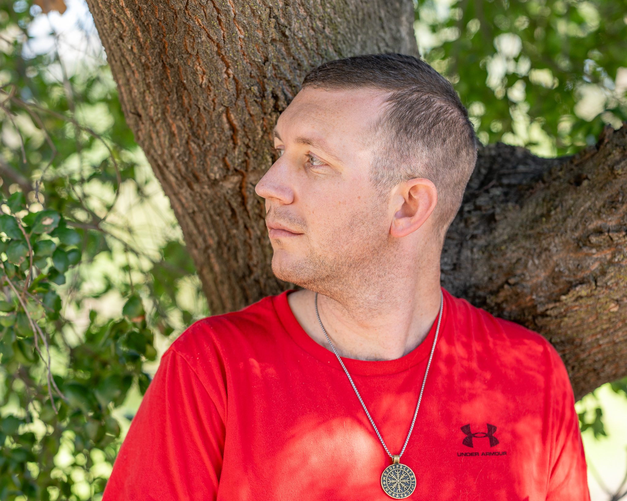 A photo of a young man leaning against a tree trunk with blurred foliage in the background. He has a short hair cut, brown hair, is wearing a red shirt with a black under armor logo. He is looking towards the left of the screen.