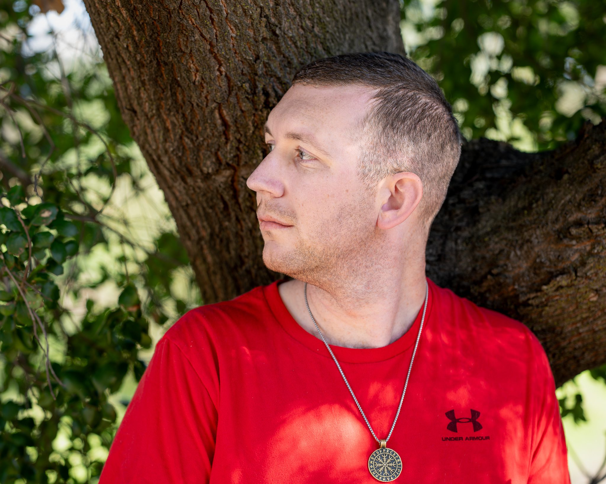 A touched up image of a young man looking towards the left of the screen. He is leaning against a tree, has short, dark blonde hair, and is wearing a red shirt with a black under armor logo on the right. The background is foliage.