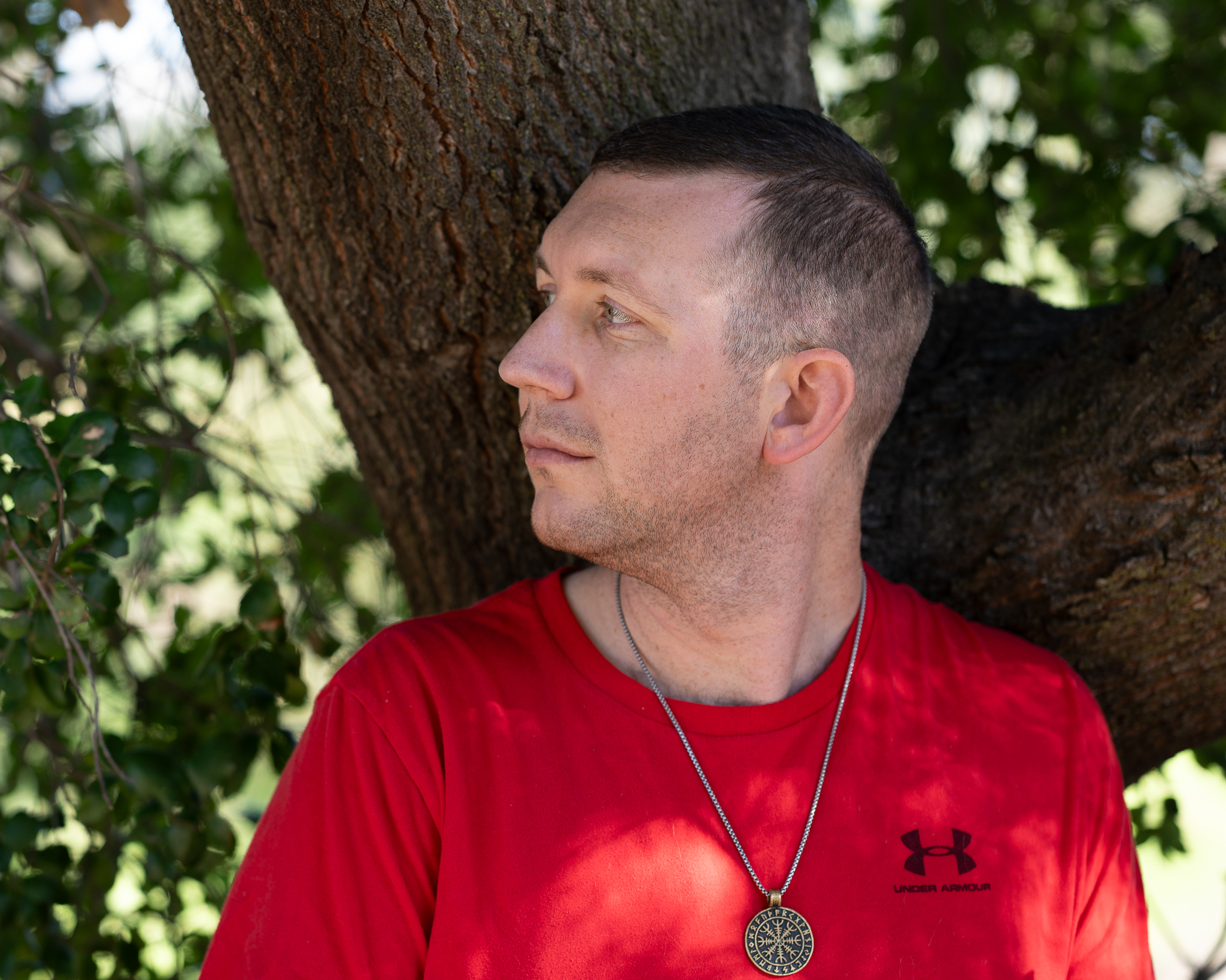 An unedited image of a young man with short dark blonde hair wearing a red shirt with a black under armor logo. He's leaning against a tree, facing the left of the screen with green blurred foliage in the back.